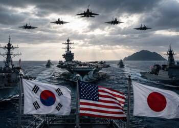 South Korean, US and Japanese flags fly aboard warships as a multinational naval fleet, including an aircraft carrier and destroyers, conducts operations at sea beneath fighter jets and dramatic storm clouds.