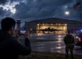 A man photographs a stealth bomber inside a brightly lit hangar from outside a barbed-wire fence, while a security officer and police vehicle stand nearby under a stormy evening sky.