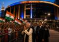 Mohan Bhagwat greets supporters with folded hands outside a brightly lit arena in New York, as Indian-American attendees wave Indian flags and security personnel escort him.