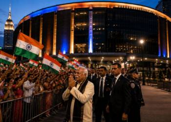 Mohan Bhagwat greets supporters with folded hands outside a brightly lit arena in New York, as Indian-American attendees wave Indian flags and security personnel escort him.