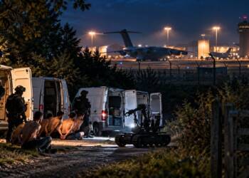 Police officers inspect three white vans with a bomb-disposal robot near a military airbase at night.” Terror