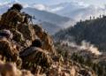 Armed fighters take positions on a rocky mountain ridge overlooking a smoke-filled Afghan valley, with distant mountains, villages and a Pakistani flag visible in the background.
