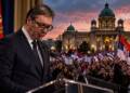 Serbian President Aleksandar Vučić resigns stands at a lectern before a large crowd waving Serbian flags, with a government building illuminated at dusk in the background.