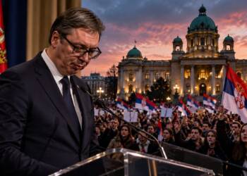 Serbian President Aleksandar Vučić resigns stands at a lectern before a large crowd waving Serbian flags, with a government building illuminated at dusk in the background.