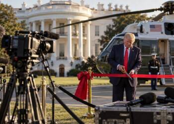 US President Donald Trump cuts a red ribbon outside the White House as television cameras and microphones sit unused, with Marine One in the background, symbolising a media blackout.