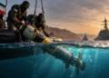 Iranian forces recover an unmanned underwater drone from the Strait of Hormuz as naval vessels patrol in the background.