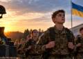 Young Ukrainian cadets in military uniforms march past a memorial for fallen soldiers at sunset, with the Ukrain flag in the background.