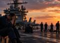 A distressed US Navy sailor sits on an aircraft carrier flight deck at sunset, with fighter jets, fellow sailors and the carrier’s island structure visible in the background.