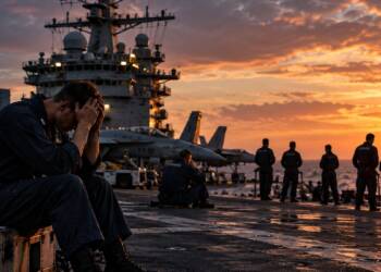 A distressed US Navy sailor sits on an aircraft carrier flight deck at sunset, with fighter jets, fellow sailors and the carrier’s island structure visible in the background.